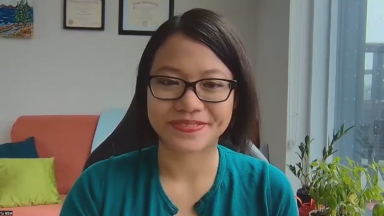 A woman with black hair, wearing dark-rimmed eyeglasses and a teal-coloured sweater, sits in an office near a window.