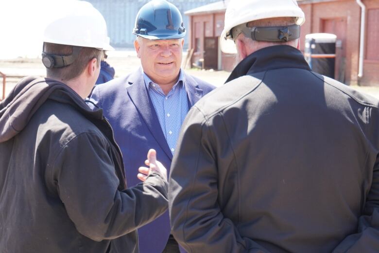 Doug Ford, wearing a suit and a hard hat, talks to two other men wearing hard hats and work clothes. 