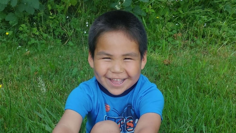 A boy in a blue shirt sits in grass and smiles at the camera.