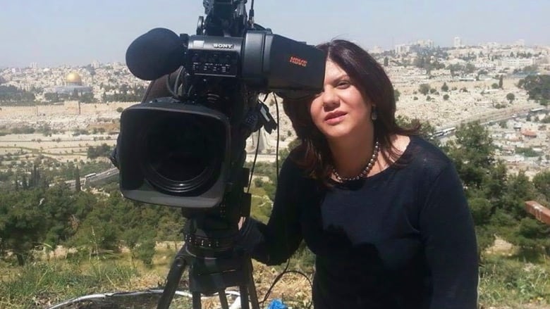 A woman wearing black looks through a TV camera in front of a background of hills and white buidlings.