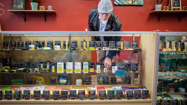 A man in black jacket and grey hat reaches down behind a counter full of cannabis products.