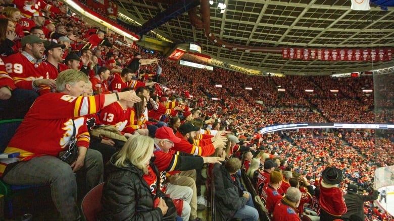 A large crowd of fans mostly dressed in red Calgary Flames jerseys are cheering in their seats at the Calgary Saddledome arena.