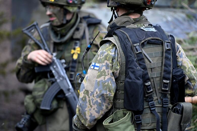 Two men in military camouflage stand, one holding an assault rifle.