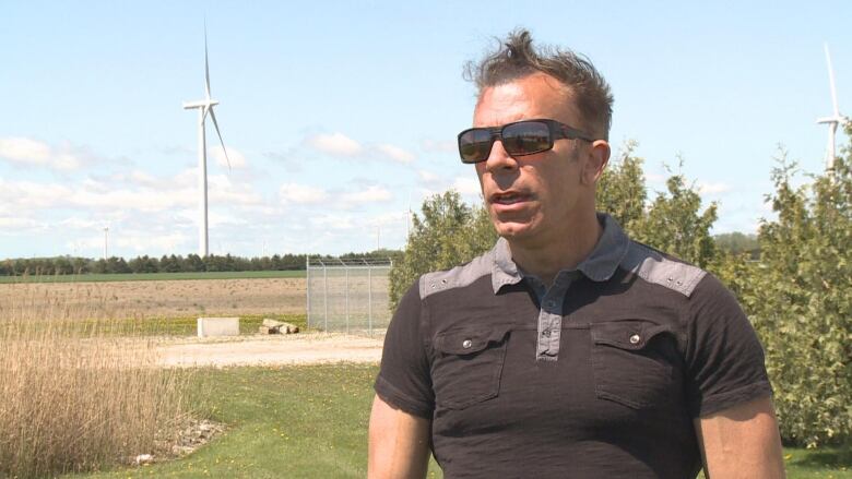 A man wearing sunglasses stands in front a field with a wind turbine in the background.