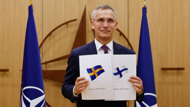 A bespectacled man in a suit holds up two documents showing the Swedish and Finnish flags.