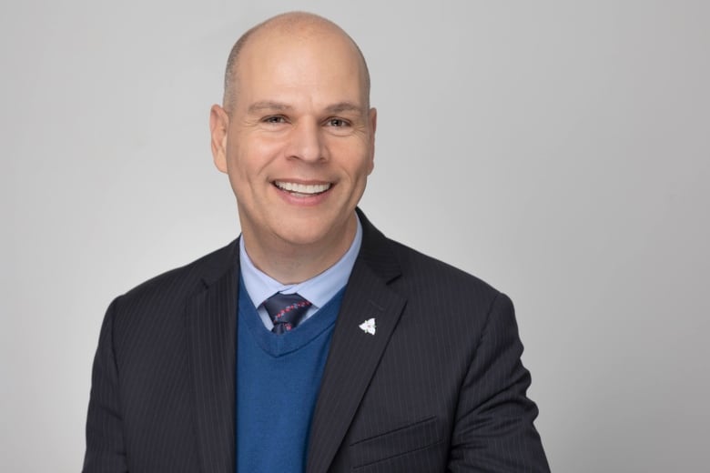 A smiling bald man wearing a navy blazer and blue vest in front of a grey background