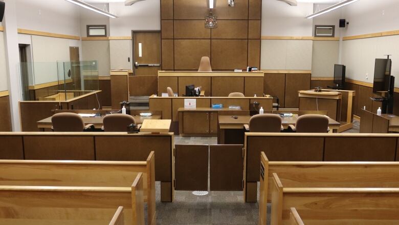 An empty court room shows the judge's bench in the distance, with a witness box and lawyers' tables nearby. There are several rows of public benches. 