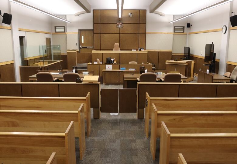 An empty court room shows the judge's bench in the distance, with a witness box and lawyers' tables nearby. There are several rows of public benches. 