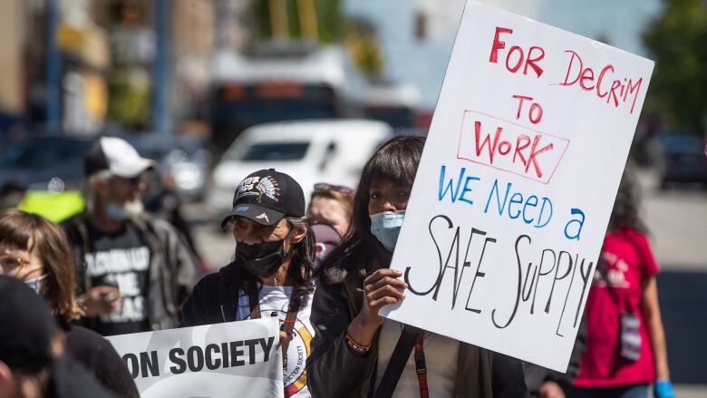 A woman wearing a mask holds up a sign that reads 'For decrim to work, we need a safe supply'. She is at the head of a row of people marching on a street.