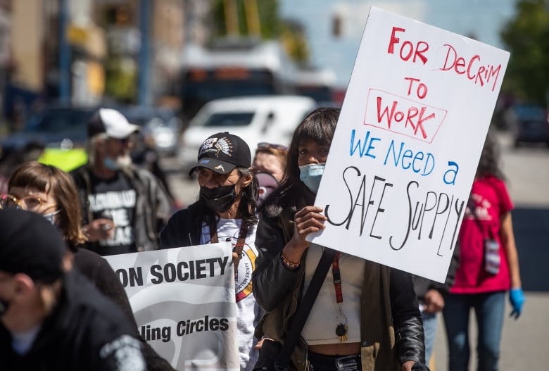 A woman wearing a mask holds up a sign that reads 'For decrim to work, we need a safe supply'. She is at the head of a row of people marching on a street.