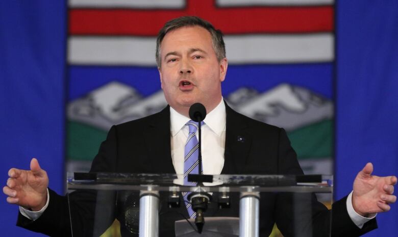 A man wearing a suit gives a speech with the Alberta flag in the background.