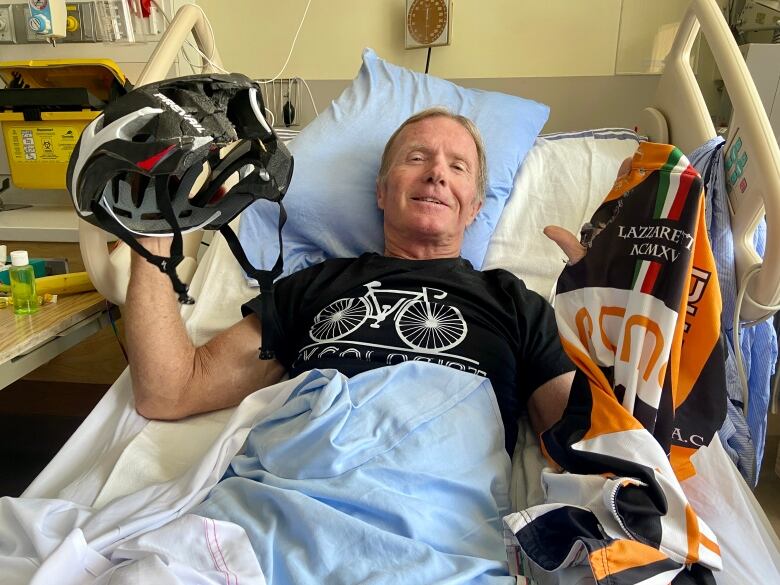 Brian Atkinson lies in a hospital bed while holding up the helmet and the jersey he was wearing when he was hit by a driver.