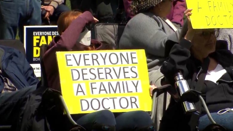 People sitting down with masks on hold a sign that says, Everyone Deserves a Family Doctor.