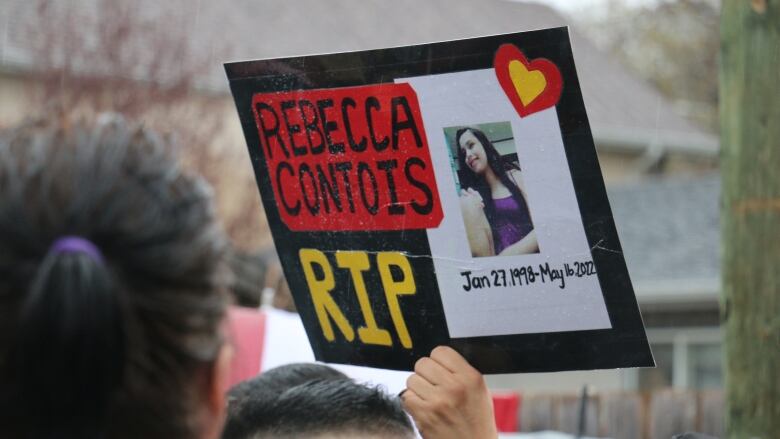 A person's hand is seen holding up a sign reading 'Rebecca Contois RIP.'