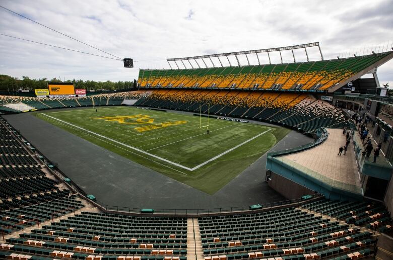 An empty football stadium sits under a cloudy sky. The seats are coloured grey, gold and green. The field features a large team logo.