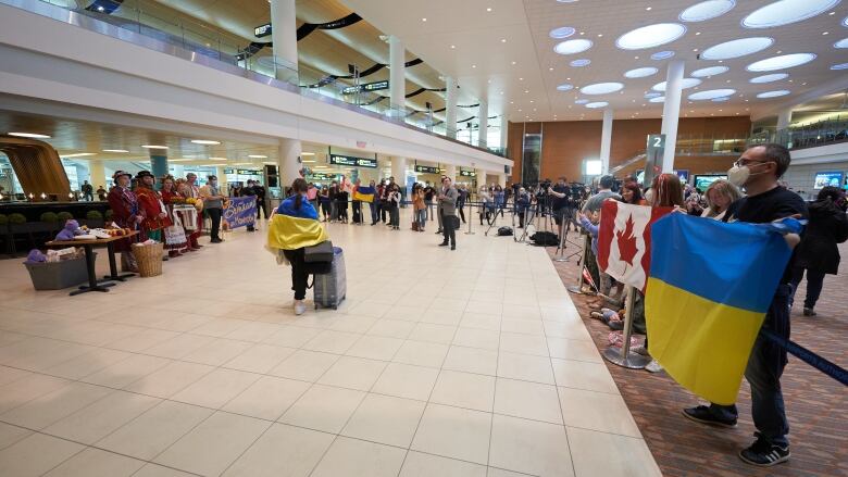 A person with a rolling suitcase and a Ukrainian flag draped over their shoulders walks past people with Ukrainian and Canadian flags at an airport.