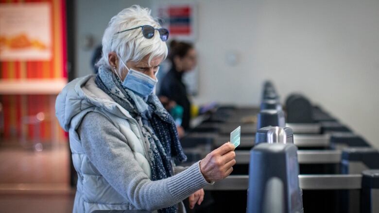 An old person taps on a fare gate at a train station. They are wearing a facemask and have short white hair.