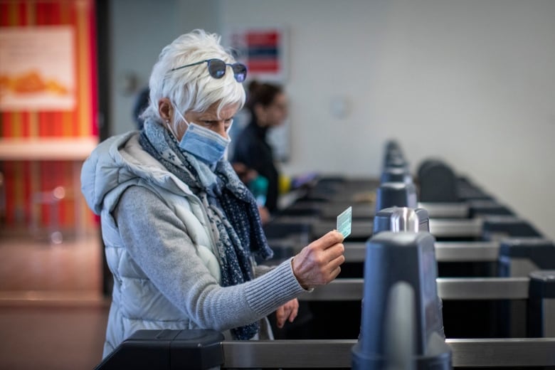 An old person taps on a fare gate at a train station. They are wearing a facemask and have short white hair.