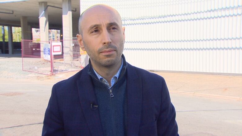 A bald man in a blue blazer and sweater stands in front of a construction site