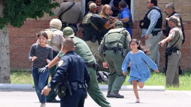 Frightened children flee a building, guided by police officers. 