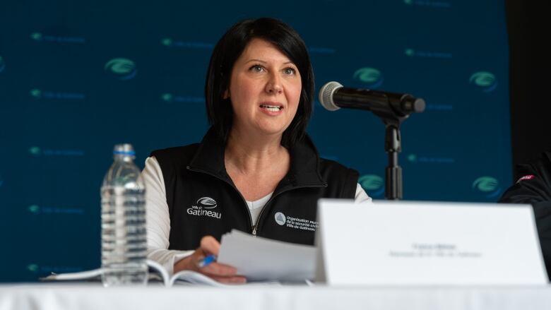 A woman sits holds papers while sitting at a table and speaking into a microphone.