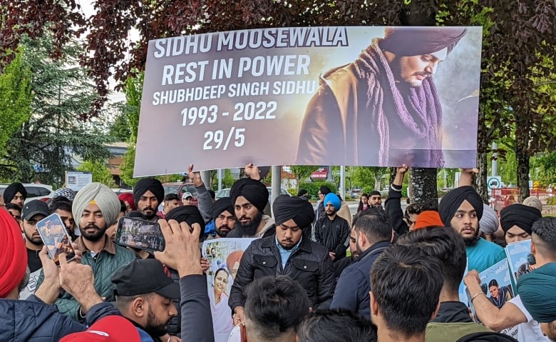 A solemn group of people gather. A large banner is held aloft that reads Sidhu Moose Wala, Rest in Power. 