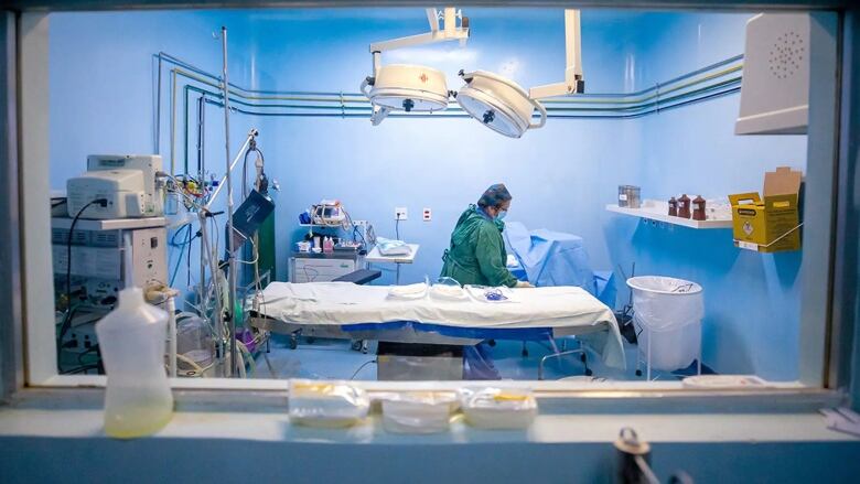 Looking through a window into an operating room where a lone health care worker, dressed in scrubs and a gown, leans on the OR table.