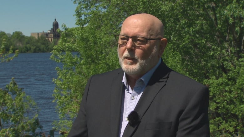 A bald man with glasses sits outside in front of green trees.