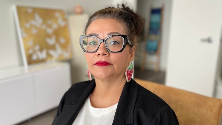 A woman in glasses, wearing strawberry-shaped earrings, looks at the camera.