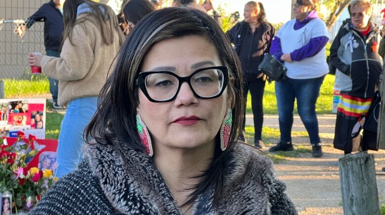 A woman with long dark hair wears bold, black-framed glasses and beaded earrings. She looks at the camera as other people stand in the background.