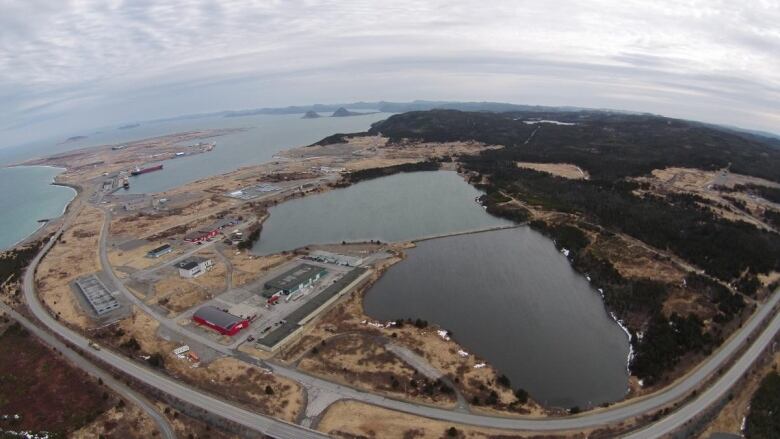 Aerial view of a port with industrial buildings mostly on the left side of the water and trees on the right.