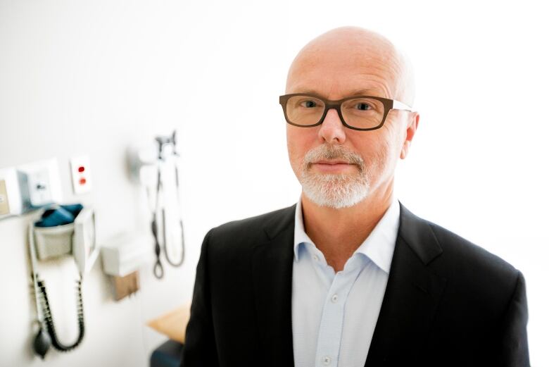 Blading man in glasses wearing a black jacket and open-neck white dress shirt, standing in a medical office.