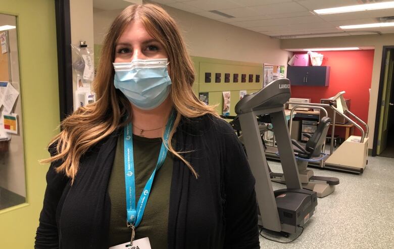 A woman in a medical mask stands in a room with exercise equipment behind her.