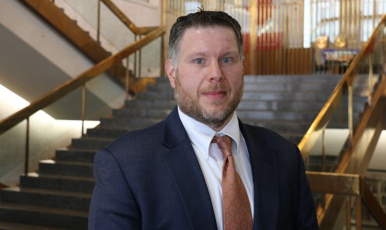 A man wears a suit and tie standing near a staircase. 