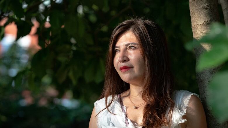 A woman with long brown hair and bright red lipstick stands next to a tree and stares off to one side with a dreamy expression. The sun through the tree leaves is reflected on her face. 