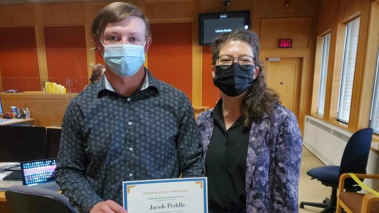 Jacob Peddle, a young man with dirty blond hair, stands in a courtroom beside a female federal crown prosecutor wearing a dark purple blazer. Both are wearing masks, with Peddle holding up the certificate of completion for graduating the Lethbridge Drug Treatment Court program. 