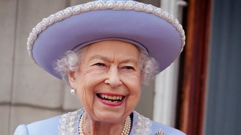 A grey-haired woman wearing a purple jacket and hat smiles.