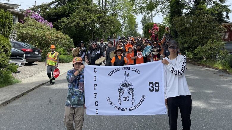 Marchers are seen coming towards the camera, with one man in a high-vis vest behind them. They are holding up a banner that reads: 