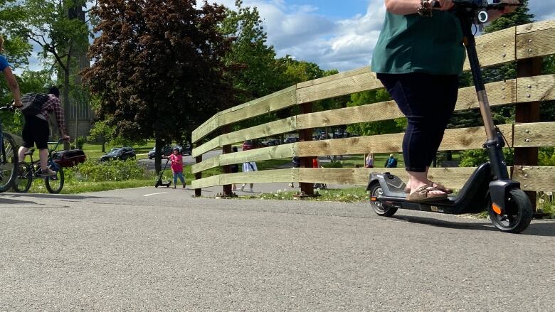 A trail with a biker on the far left and a e-scooter rider in the foreground on the right.