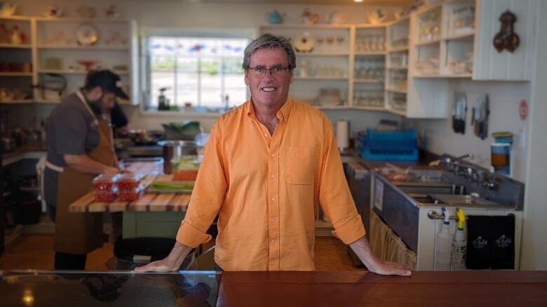 Derrick Hoare stands at a table smiling in an orange shirt as a chef works behind him in the kitchen. 