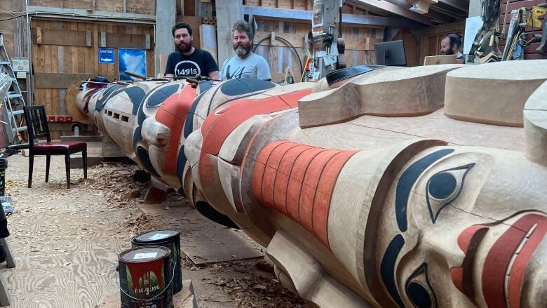 A large totem pole in progress lies on its side with two brothers standing near the head of the pole.