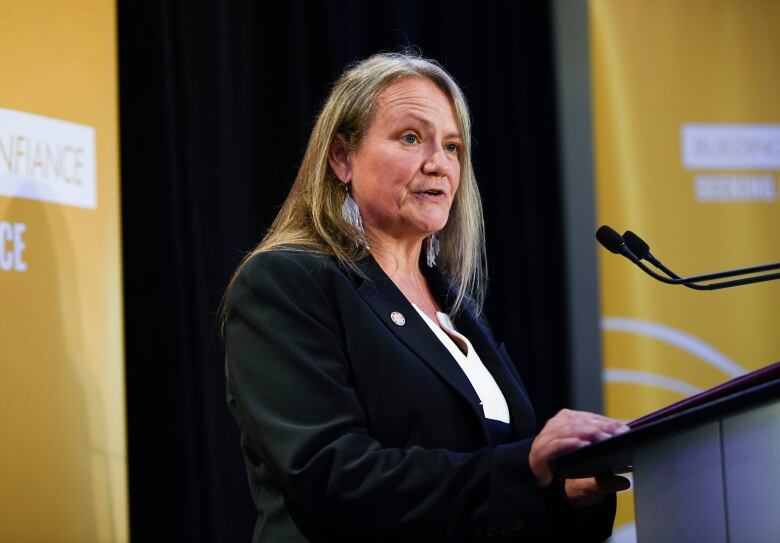 A woman with grey hair wearing a suit and beaded earrings speaks at a podium