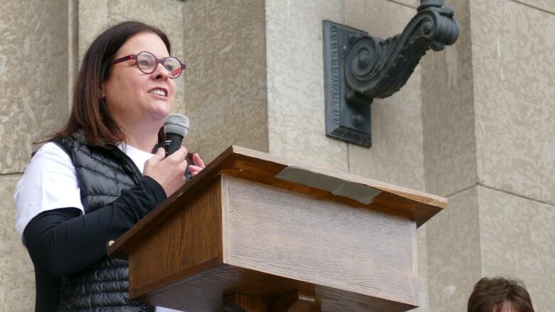 A woman in glasses and a vest holds a microphone while standing at a podium on the steps of the legislature.