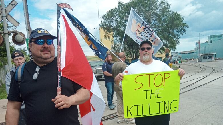 Workers wave flags outside a factory. One holds a bristol board sign that says 