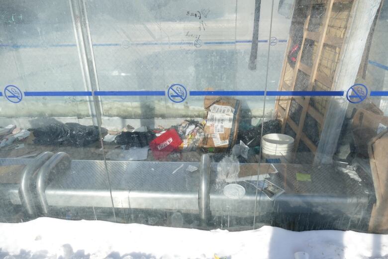 Garbage, including tattered clothes, black garbage bags, boxes and coffee cup lids, is strewn across a metal bench and concrete floor inside a bus shelter.