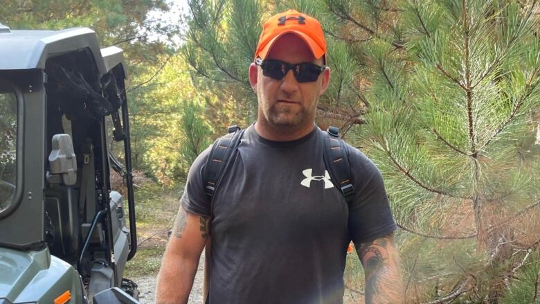 A man stands in a wooded area wearing an orange hat and sunglasses.