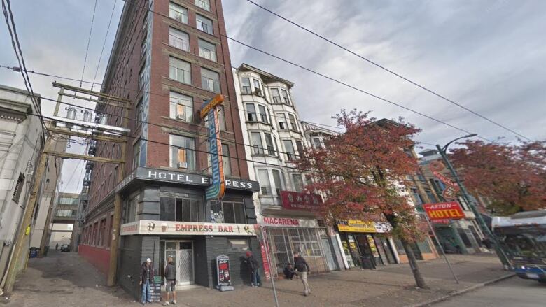 A long slim building stands next to an alley and other nearby hotels on a pavement. A sign reads 'Hotel Empress', with a smaller sign below it reading 'Empress Bar'. A few people are on the pavement. A bus is visible to the side.