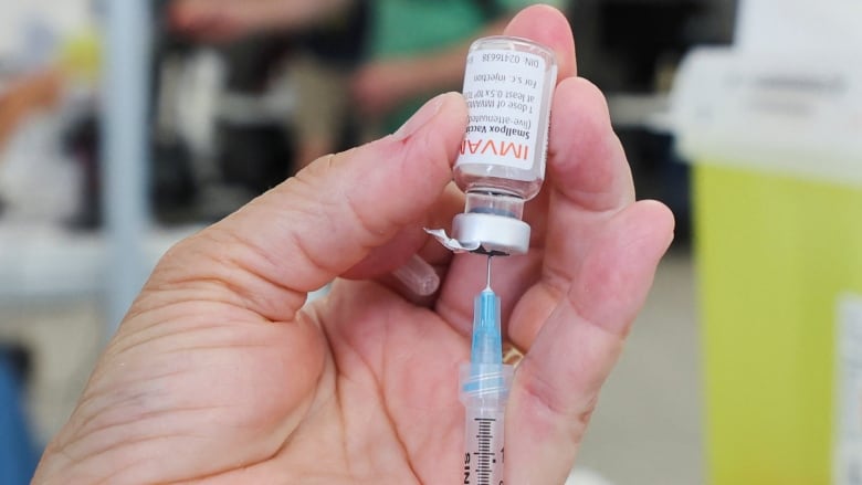 Closeup of a a hand sticking a need tip into an upside down vial of monkeypox vaccine.