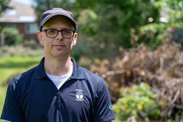 A man in a baseball cap stands in front of downed trees.