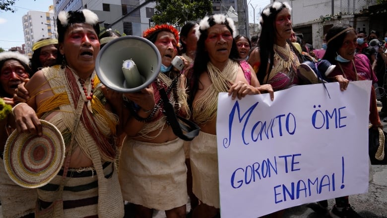 Indigenous Ecuadorians wearing face paint and traditional attire hold a protest sign.